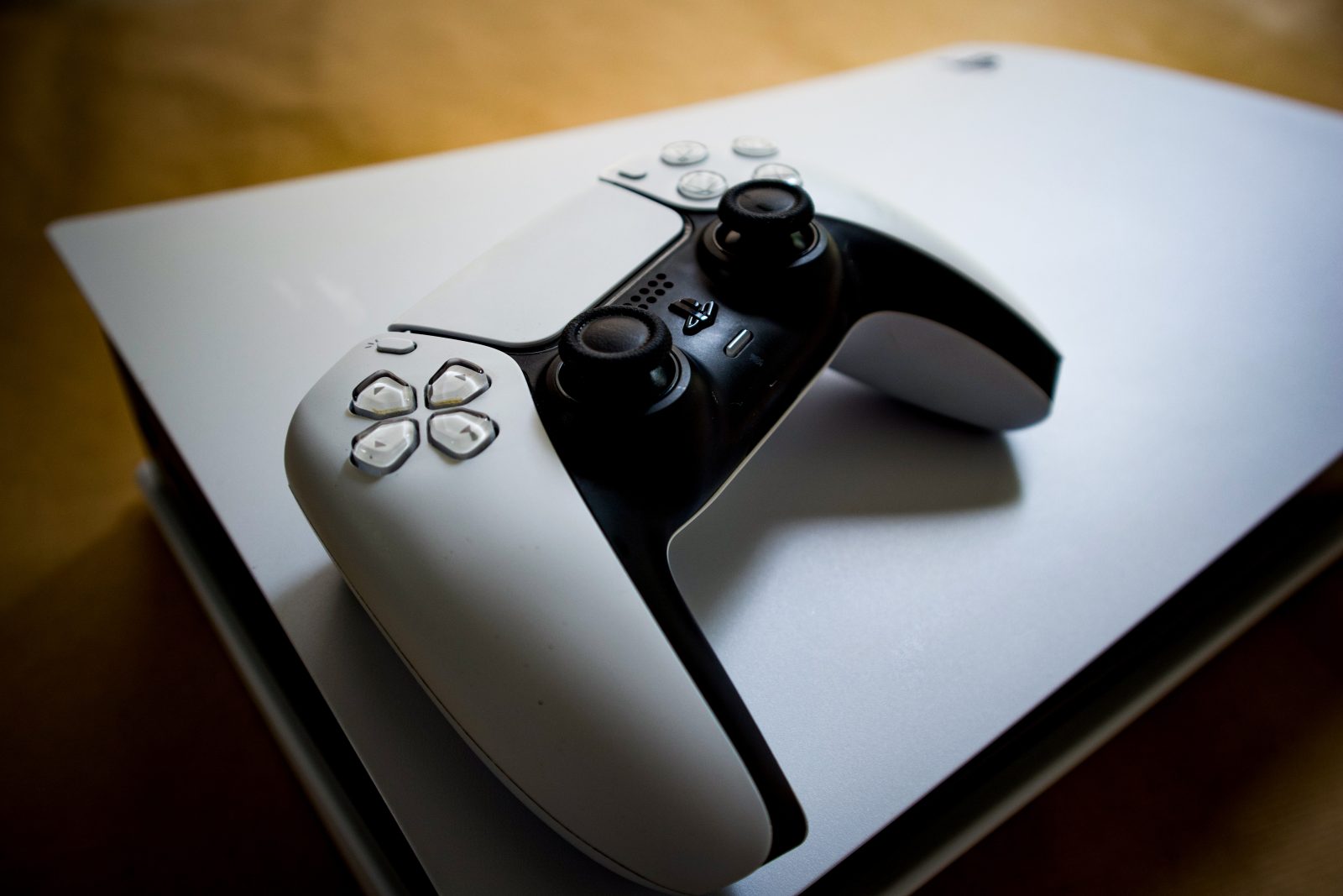 Witte PlayStation DualSense controller liggend op zijn kant bovenop een horizontaal geplaatste PS5 console, met een houten ondergrond op de achtergrond
