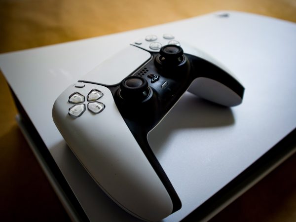 Witte PlayStation DualSense controller liggend op zijn kant bovenop een horizontaal geplaatste PS5 console, met een houten ondergrond op de achtergrond