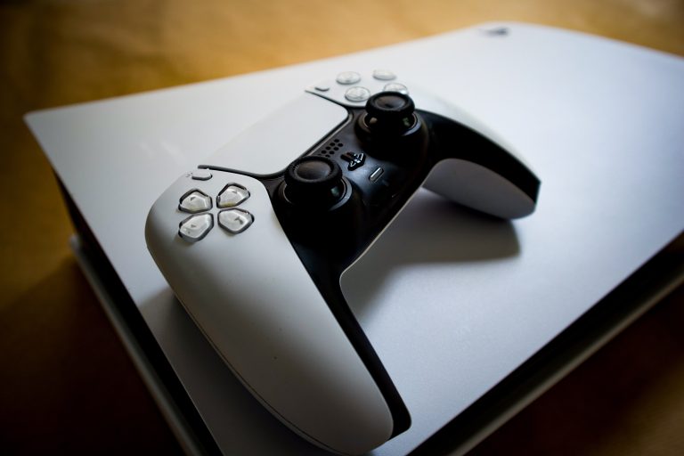 Witte PlayStation DualSense controller liggend op zijn kant bovenop een horizontaal geplaatste PS5 console, met een houten ondergrond op de achtergrond