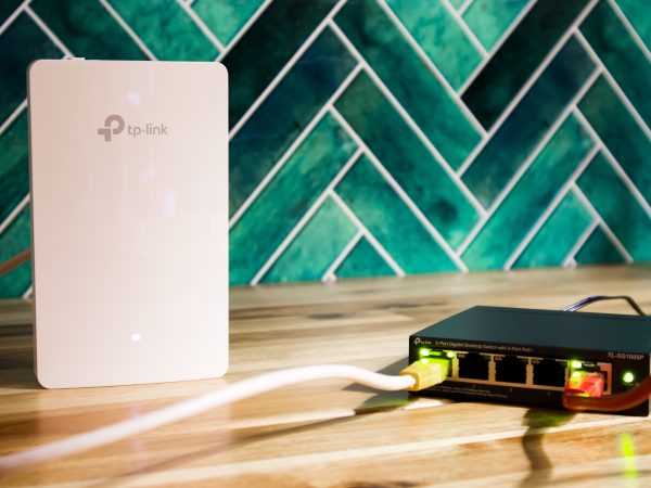 Wit TP-Link accesspoint naast een zwarte TP-Link netwerkswitch met verlichte poorten, op een houten oppervlak met een groenblauwe tegelwand op de achtergrond