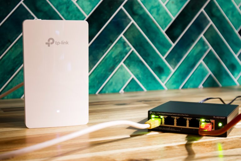 Wit TP-Link accesspoint naast een zwarte TP-Link netwerkswitch met verlichte poorten, op een houten oppervlak met een groenblauwe tegelwand op de achtergrond